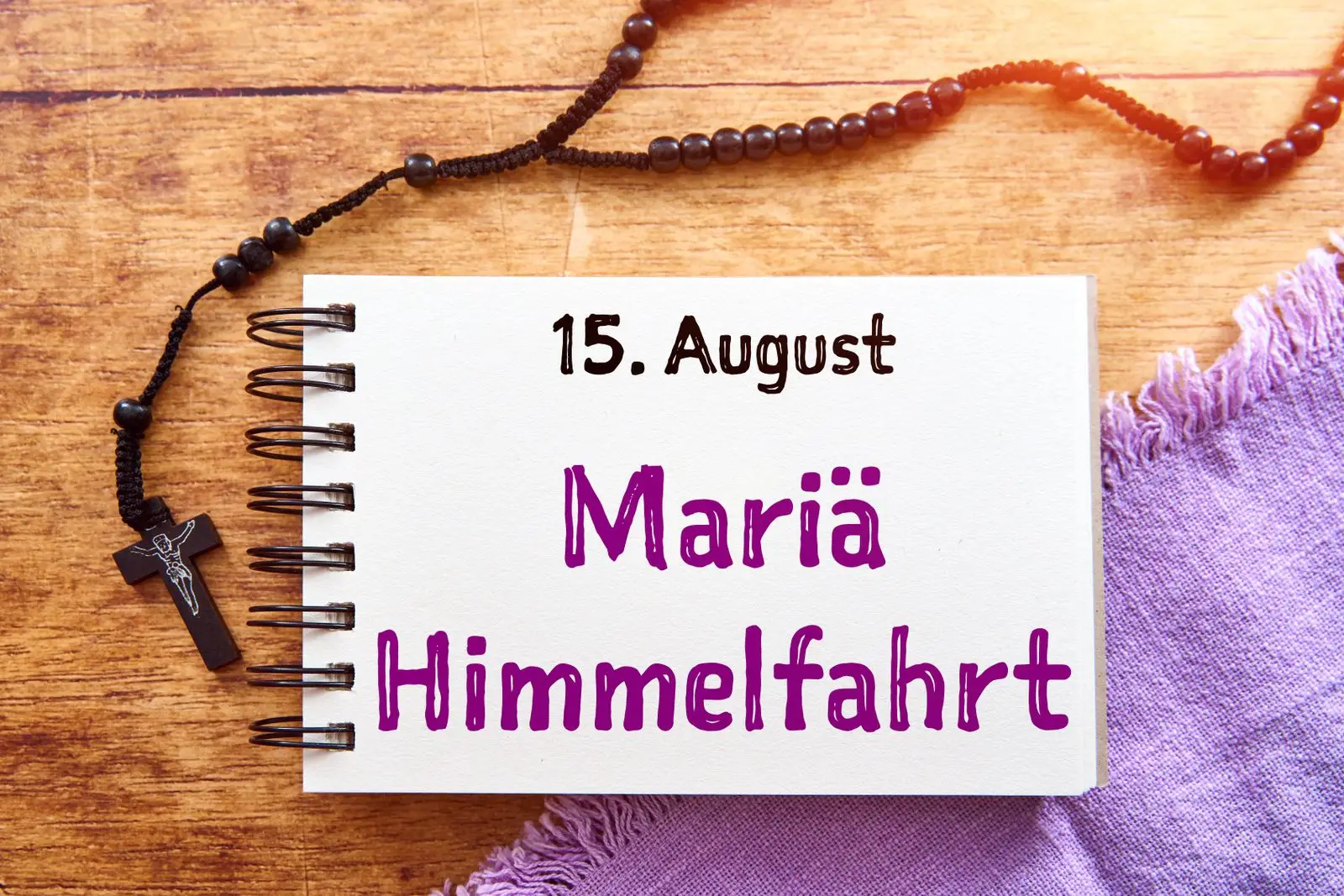 Buch mit dem text ‚15. August mariä himmelfahrt‘, daneben ein schwarzer rosenkranz auf einem holztisch. Das bild erinnert an den feiertag im sommer.
