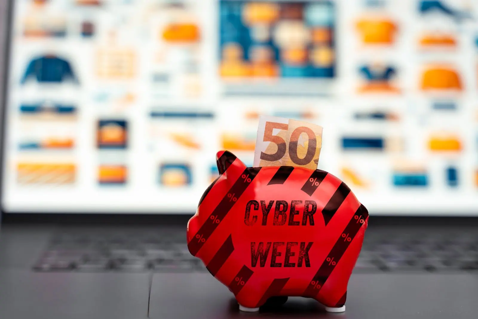Ein sparschwein mit der aufschrift ‚cyber week‘ und einem 50-euro-schein auf einem laptop. Im hintergrund unscharf eine website mit verschiedenen angeboten. Das bild symbolisiert die rabatte und sparmöglichkeiten während der cyber week und am cyber monday.