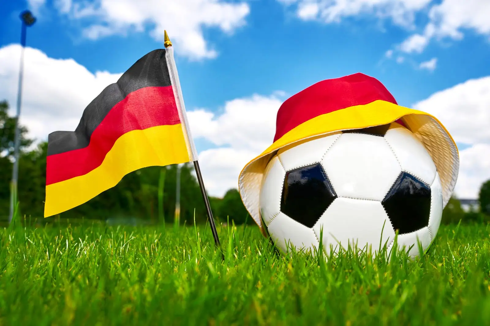 Deutschland spiel mit fußball, deutschlandflagge und fanhut in schwarz-rot-gold auf grünem rasen unter blauem himmel.