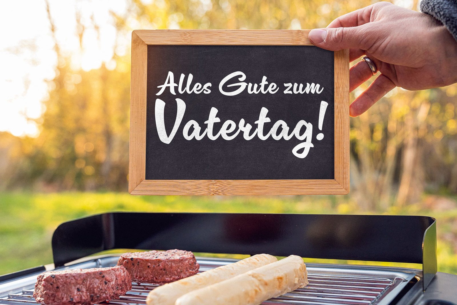 Hand hält tafel mit ‚alles gute zum vatertag‘ über einem grill mit fleisch und würstchen im freien