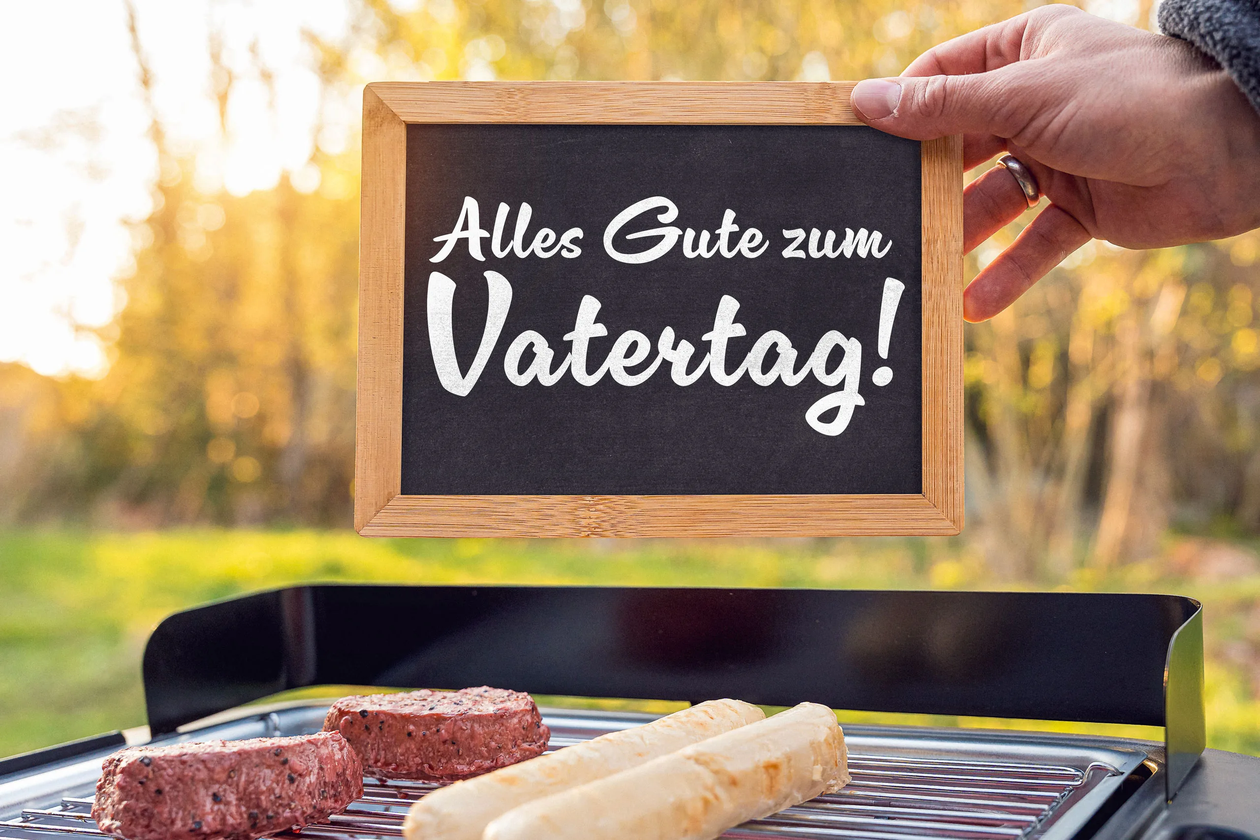 Hand hält tafel mit ‚alles gute zum vatertag‘ über einem grill mit fleisch und würstchen im freien