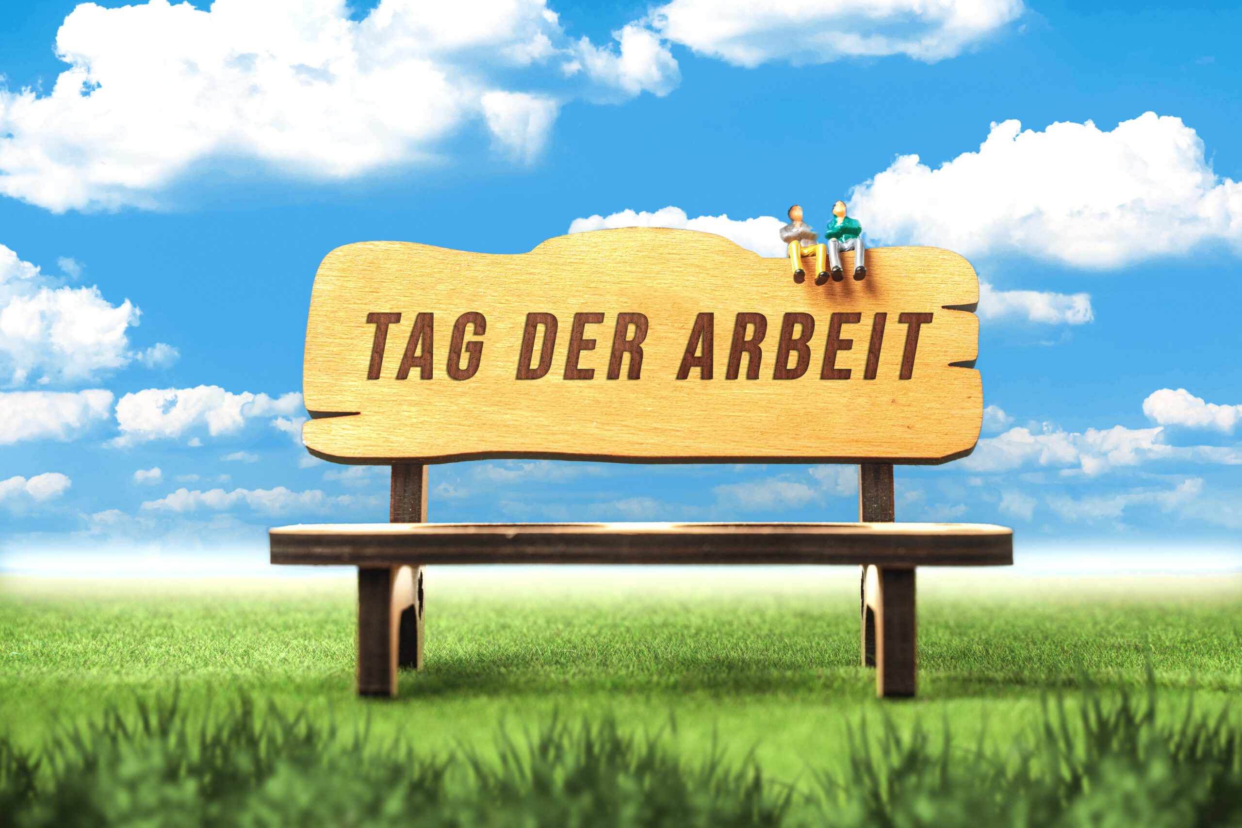 Miniatur-figuren auf einer parkbank mit der aufschrift tag der arbeit vor blauem himmel